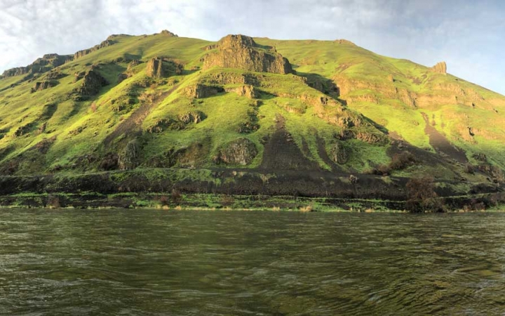 A body of water flows in front of a very green hillside